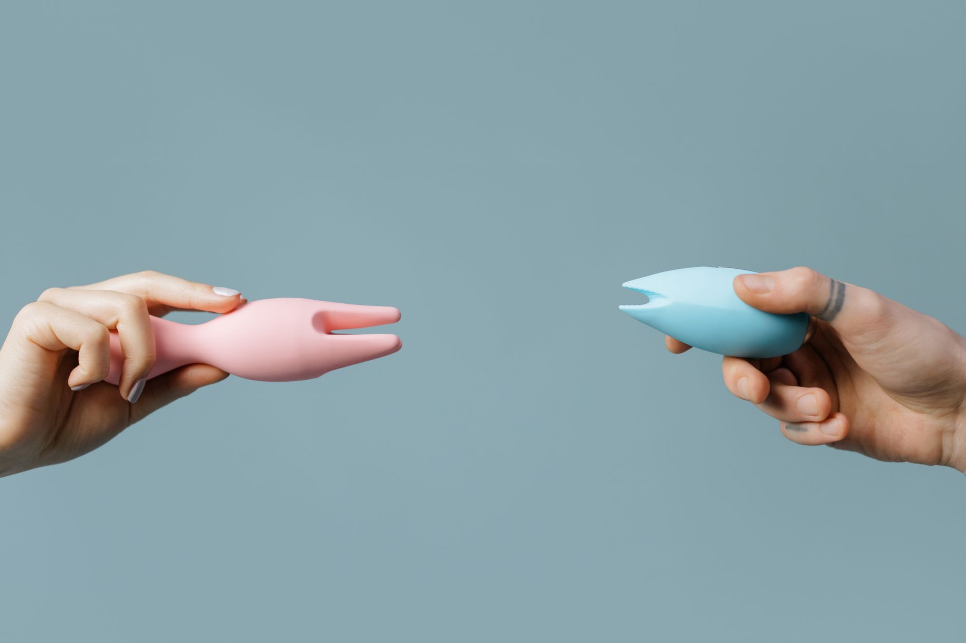 Two hands holding pink and blue silicone vibrators against a pastel background, representing pleasure tools for bridging desire gaps.